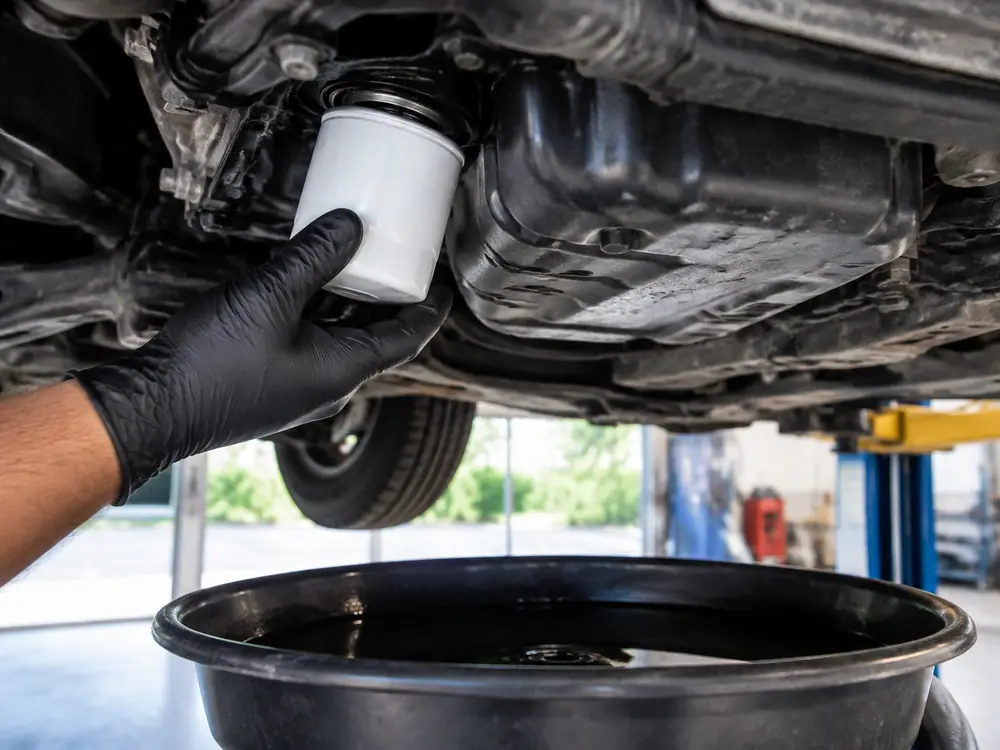 A gloved hand fitting a new oil filter under a car, with a drain pan of used oil below